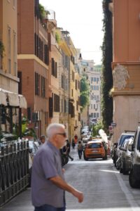 A busy street with people walking and cars parked against buildings, with pedestrians and vehicles in the background.