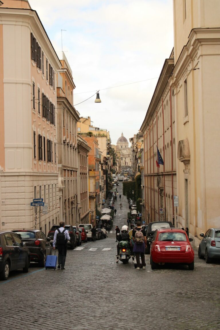 A cobblestone street lined with parked cars and people