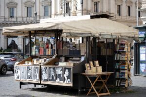 A vintage bookstall spills paperbacks, prints, and black‑and‑white portraits onto a cobbled Roman square.