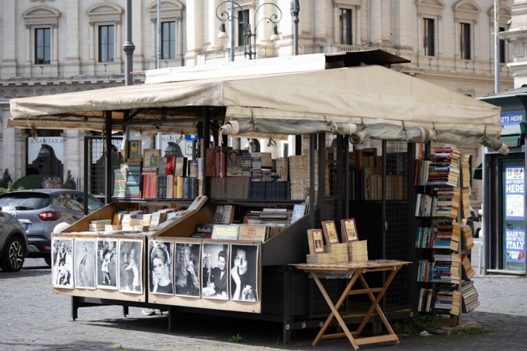 A vintage bookstall spills paperbacks, prints, and black‑and‑white portraits onto a cobbled Roman square.